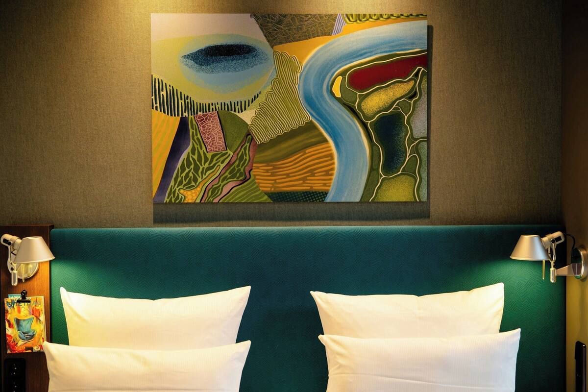 Ein Hotelzimmer mit einem großen, abstrakten Gemälde, einem türkisblauen Kopfteil und zwei weißen Kissen.