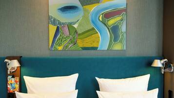 Hotelkamer met twee bedden, een blauwgroen hoofdeinde, een wanddecoratie abstract kunstwerk en een nachtkastje met een lamp.