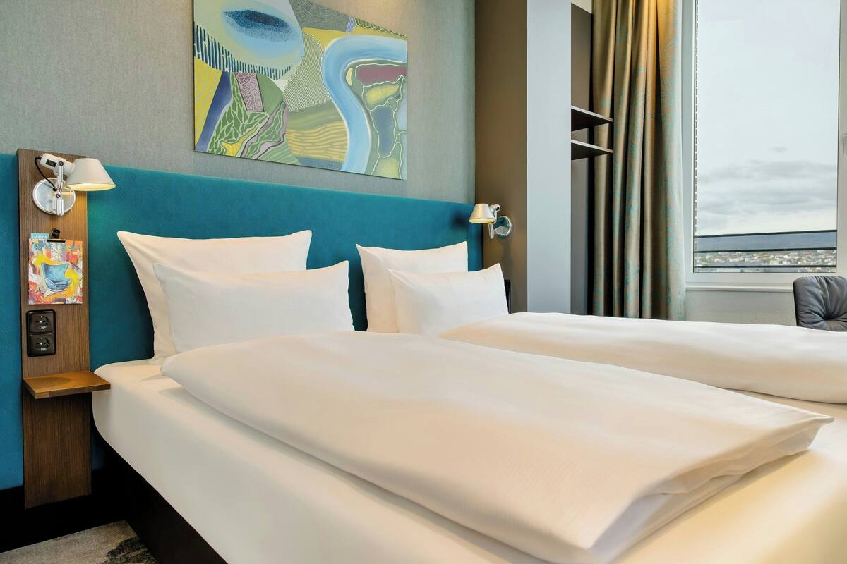 Ein Hotelzimmer mit zwei Betten, einem blauen Kopfteil, einem Bild an der Wand und Blick auf die Stadt durch das Fenster.