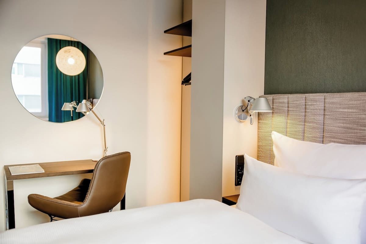 A hotel room with a bed, a desk with a chair, a round mirror, and a wall-mounted lamp.