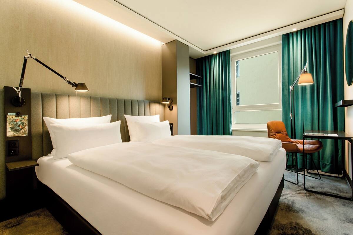 A hotel room with two beds, a desk, a chair, and a large window with curtains.
