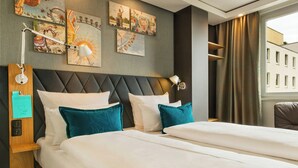 A hotel room with a large bed, two bedside tables, a headboard with a built-in lamp, artwork on the wall, and a view of a building through a window.