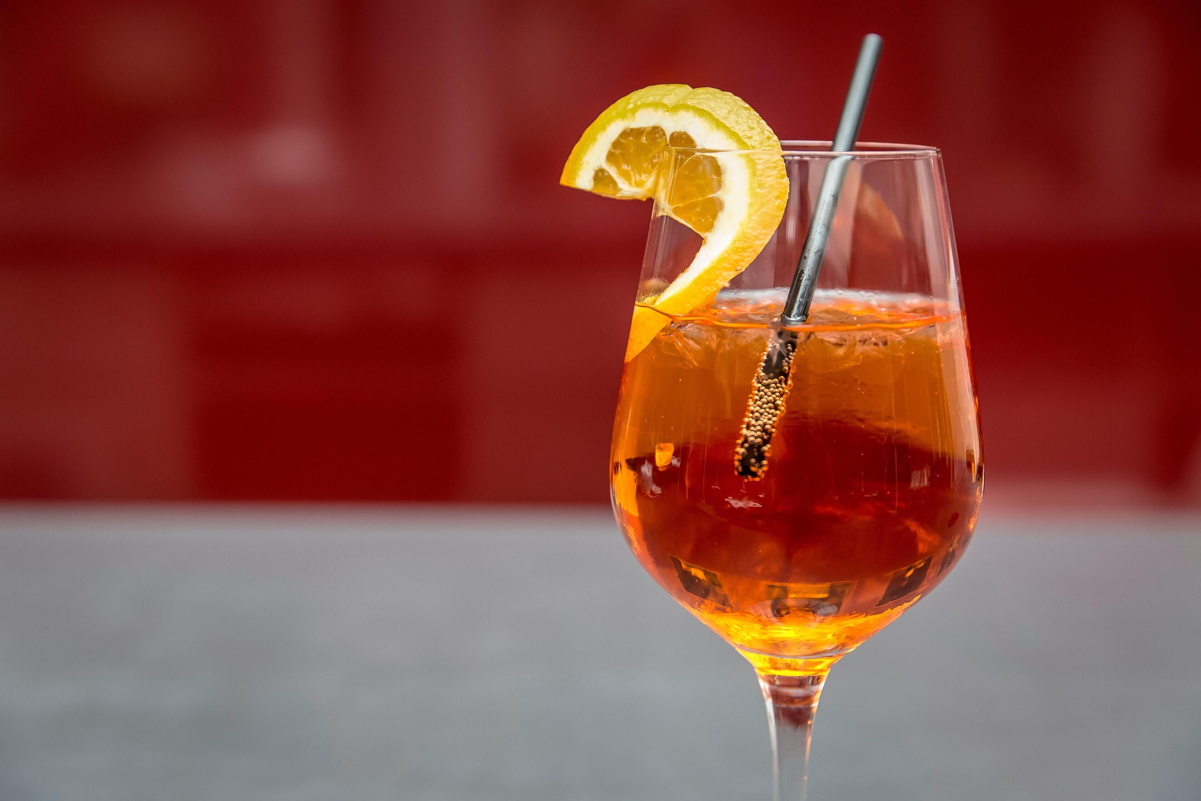 Et glas med en ravfarvet drik, et citronskive og et sugerør, foran en sløret rød baggrund.