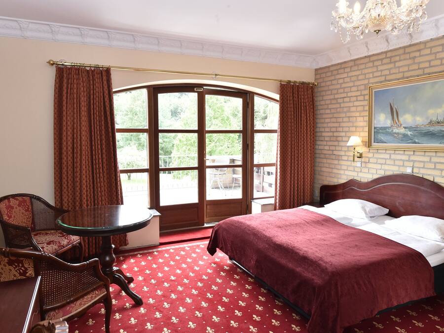 Habitación de hotel con un ventanal grande, una cama con colcha roja, una silla de madera y una mesita redonda.