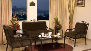 A room with a sofa, two chairs, a small table with a tray of items, and a view of the city at dusk. - Ramanashree Brunton (Bengaluru)