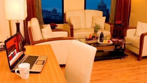 A hotel room with a sofa set, a dining table with a laptop and a mug, and a fruit basket on the coffee table. - Ramanashree Brunton (Bengaluru)