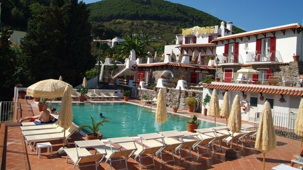 A pool area with lounge chairs and umbrellas, surrounded by buildings with red and white facades, set against a hillside backdrop. - Don Pedro Hotel (Ischia)