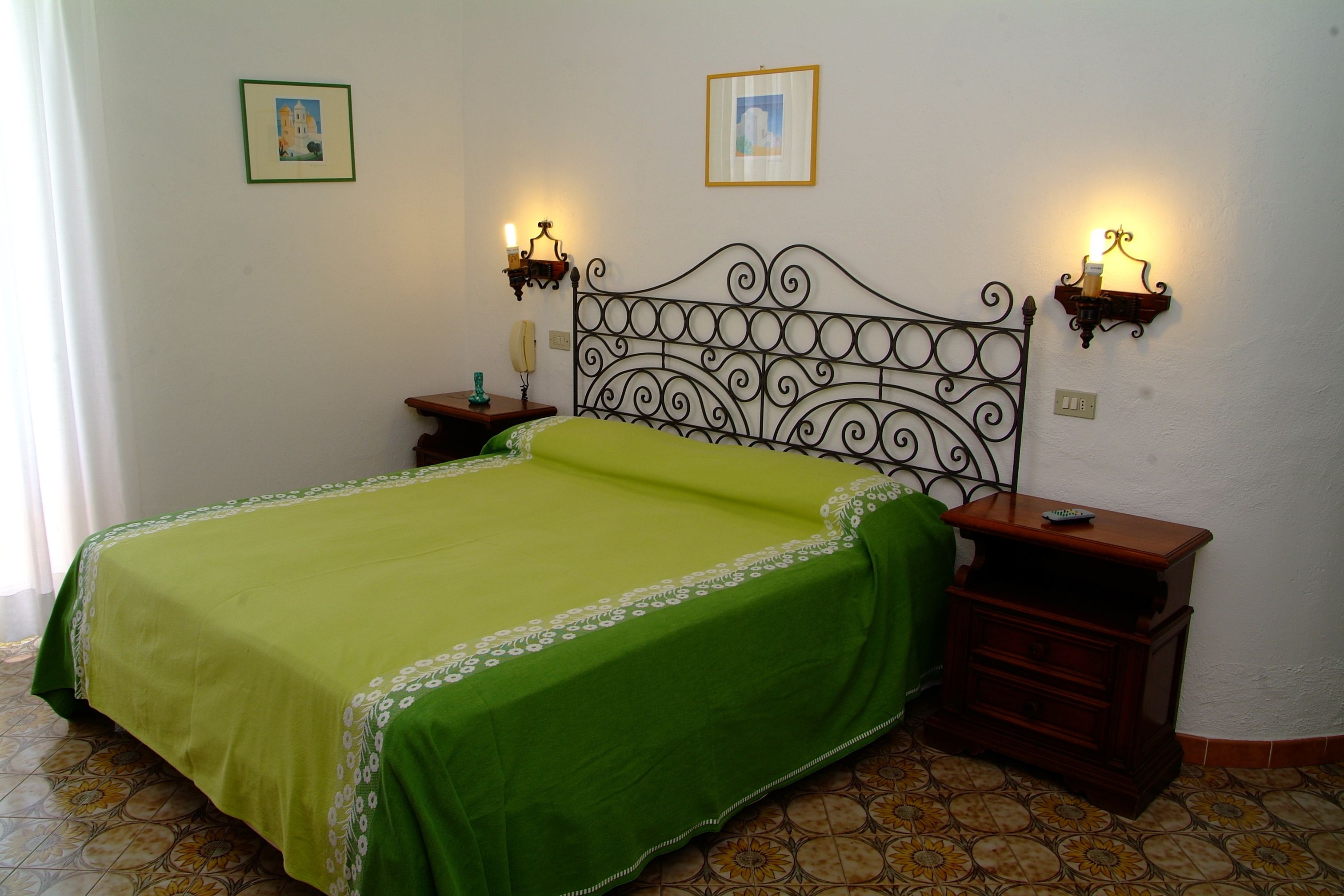 A bedroom with a green bedspread, a metal headboard, two bedside tables with lamps, and two framed pictures on the wall.