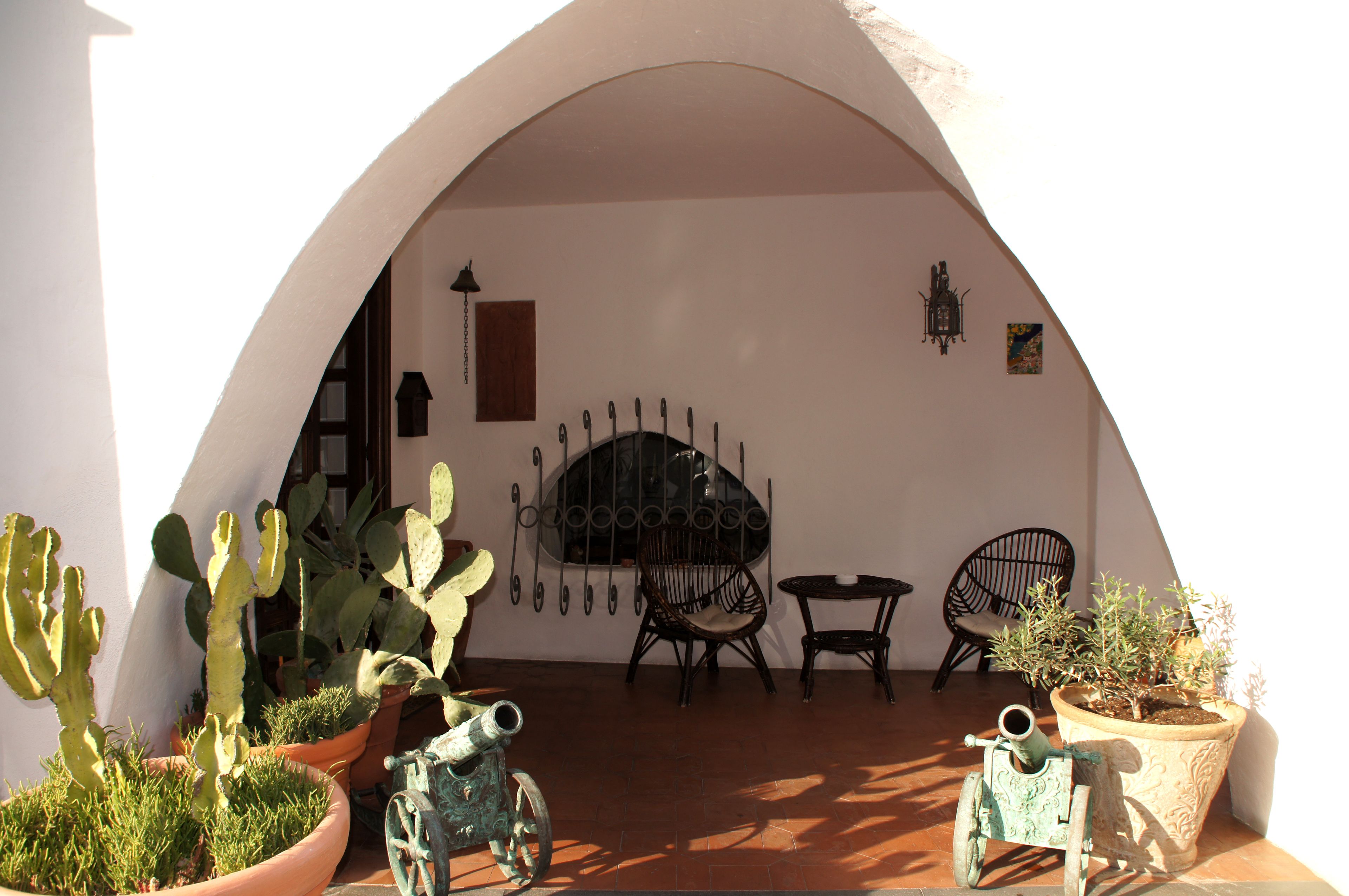 Een patio met bogen, potten met cactussen, een ijzeren hekwerk en twee rieten stoelen.