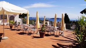 An outdoor terrace with tables and chairs, potted plants, and a view of the sea and mountains. - Don Pedro Hotel (Ischia)