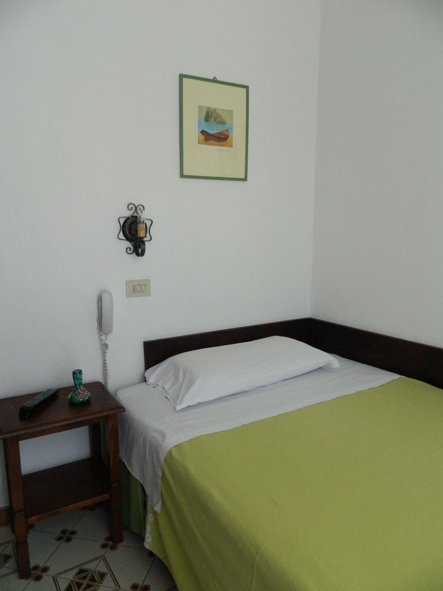 Una habitación sencilla con una cama, una mesita de noche con un teléfono, un jarrón y un cuadro en la pared.