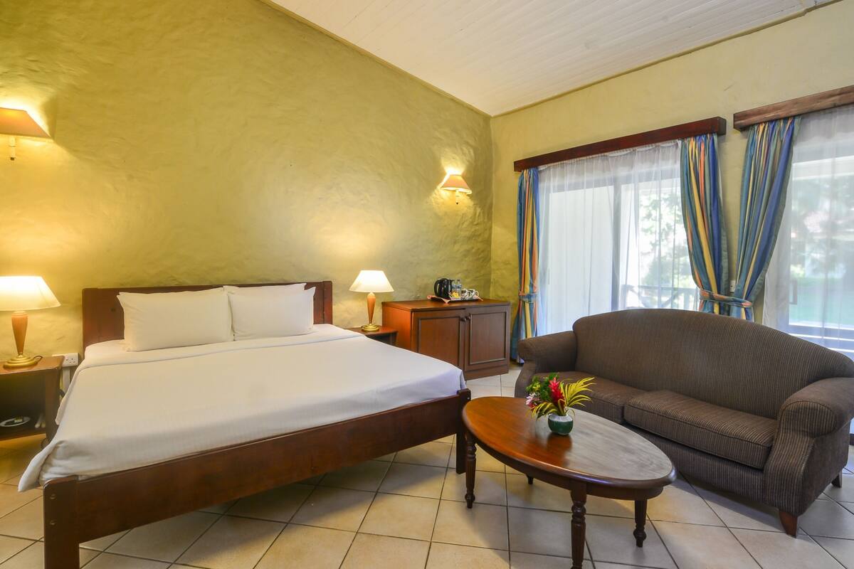 A hotel room with a bed, a sofa, a small table with a plant, a nightstand, a lamp, and a window with curtains.