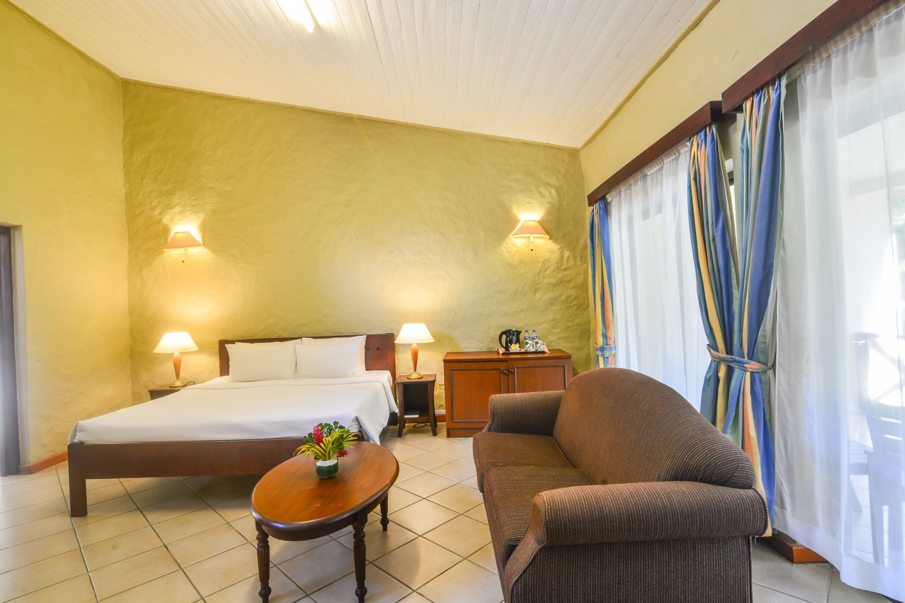 Une chambre d’hôtel comprenant un lit, un canapé, une petite table avec une plante, deux lampes de chevet, une table de nuit et un placard.