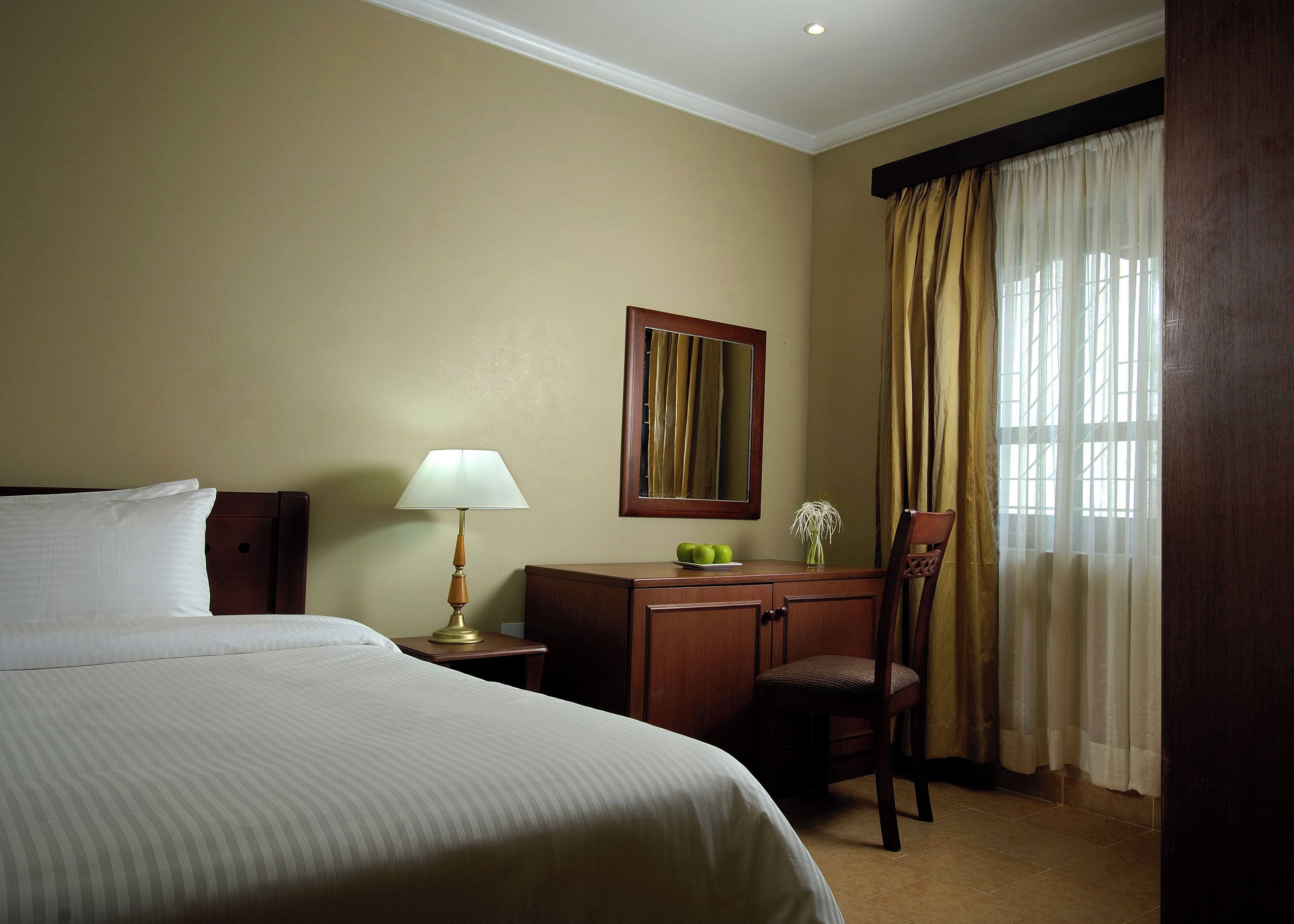 Habitación de hotel con cama, escritorio con lámpara, silla, espejo y ventana con cortinas.