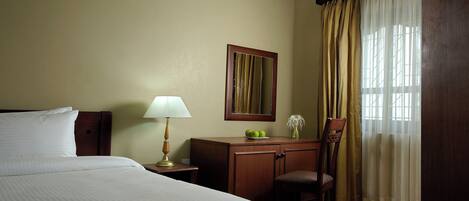 Habitación de hotel con cama, escritorio con lámpara, silla, espejo y ventana con cortinas.