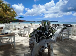 A beachside seating area with white chairs and tables, a white picket fence, and a backdrop of the sea and mountains.