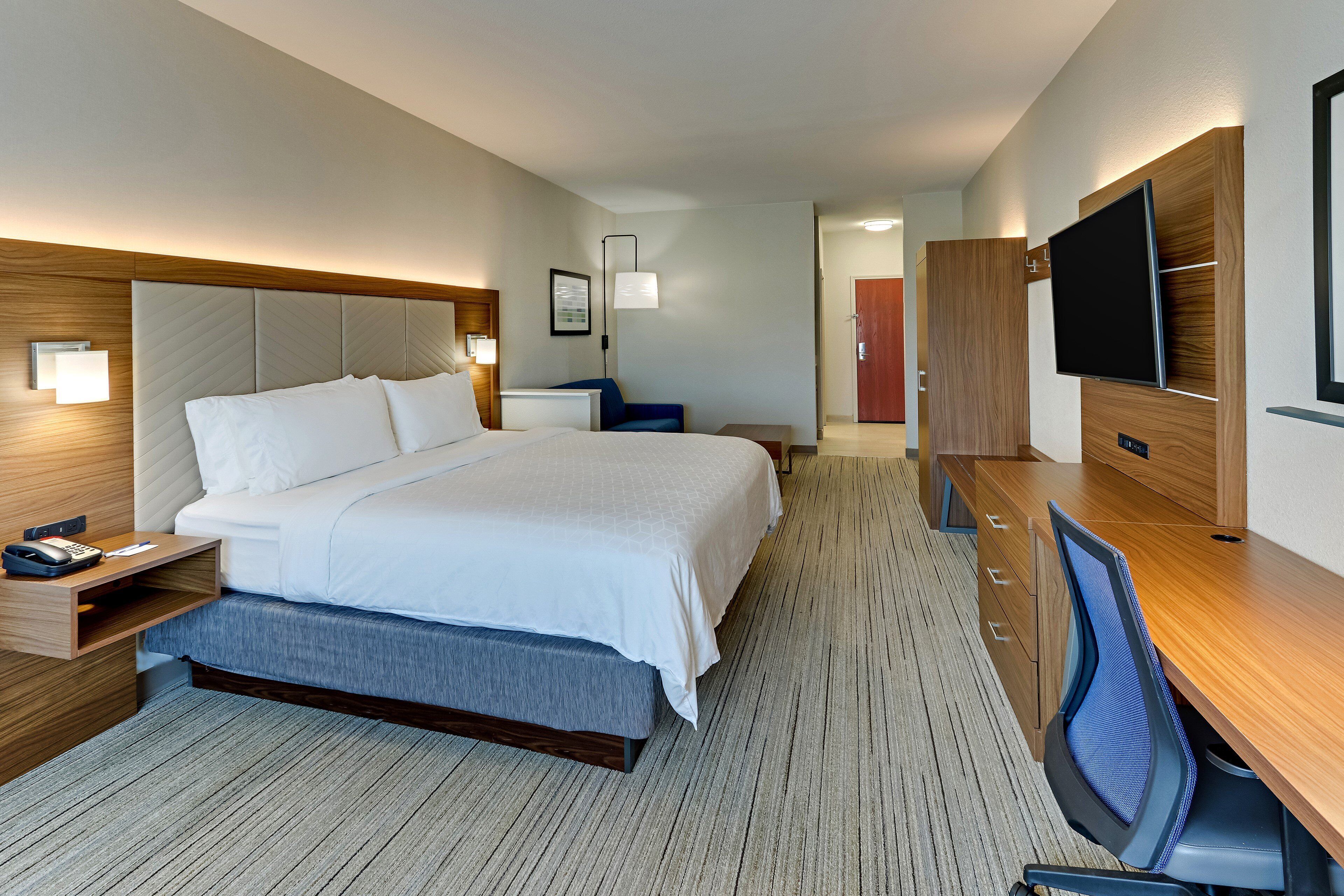 Una habitación de hotel con una cama grande, un escritorio con una silla, un televisor y una mesita de noche con lámpara.