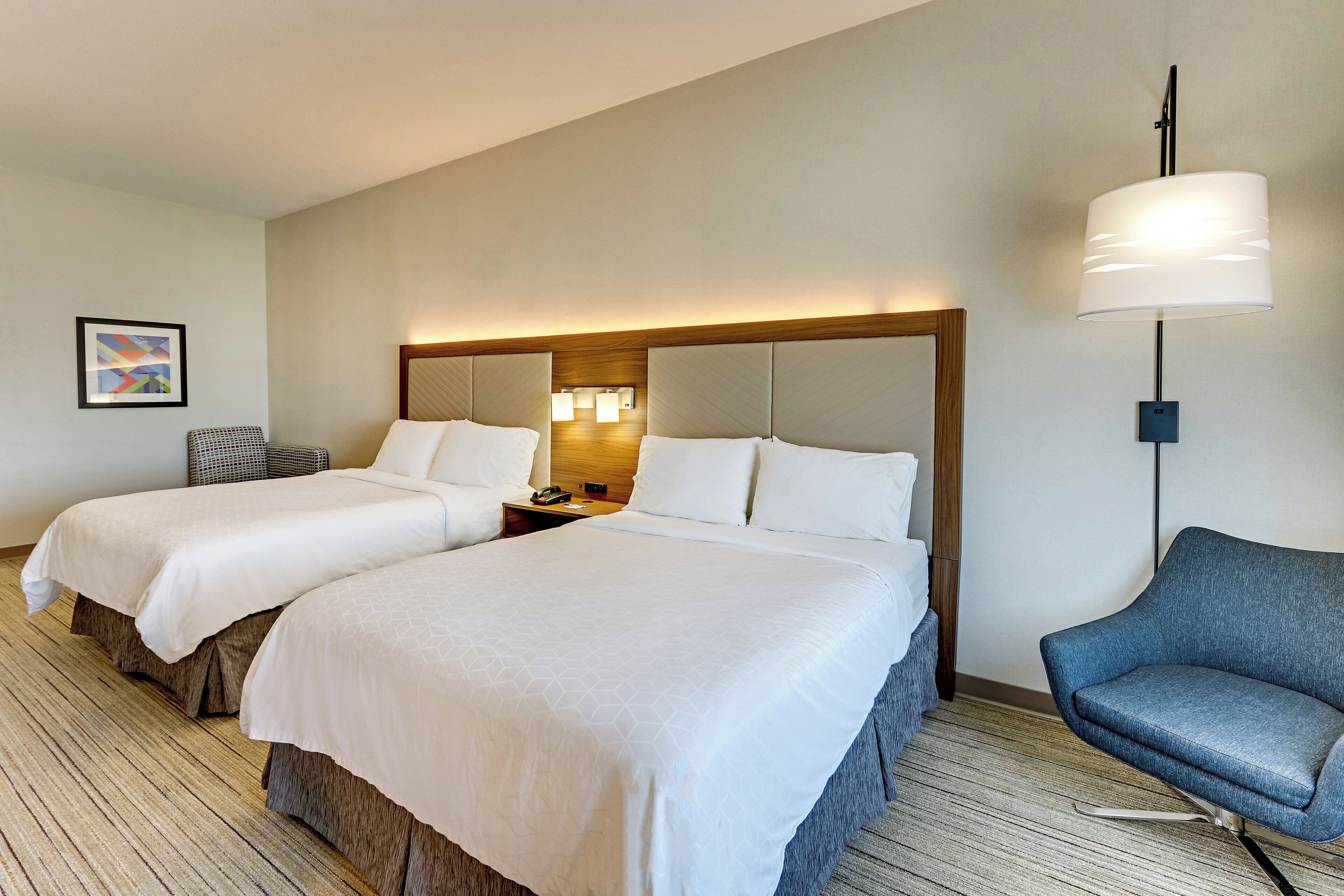 A hotel room with two beds, a nightstand with a lamp, a chair, and a framed picture on the wall.