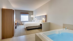 Une chambre d’hôtel avec un grand lit, un coin salon avec un fauteuil et une table, une baignoire et une fenêtre avec des stores.
