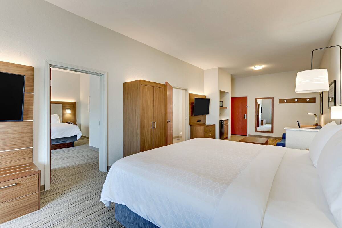 A hotel room with a large bed, wooden wardrobe, a desk with a lamp, a television, and a bathroom visible through an open door.
