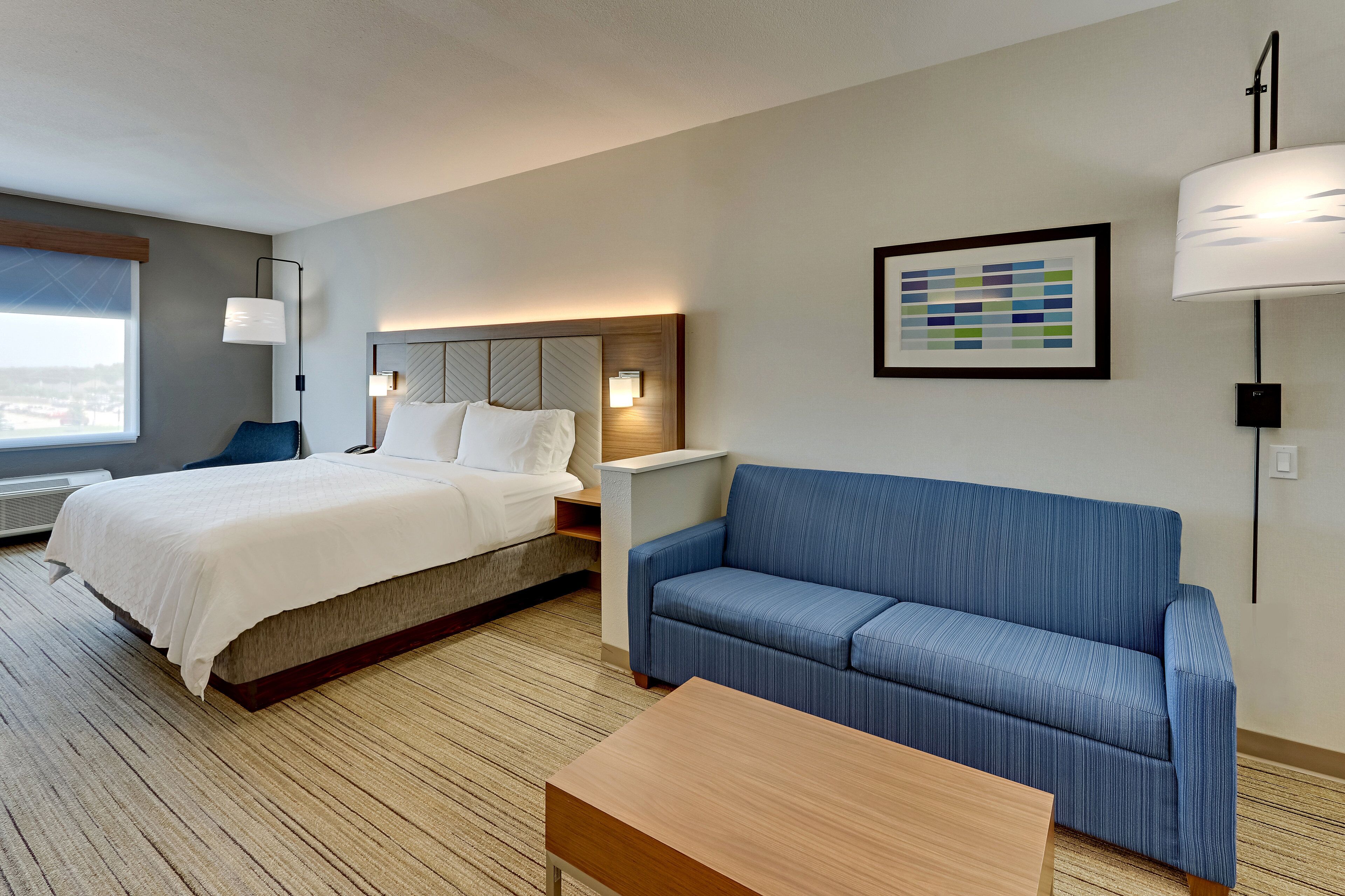 A hotel room with a bed, a blue sofa, a wooden coffee table, a nightstand, and a wall-mounted lamp.