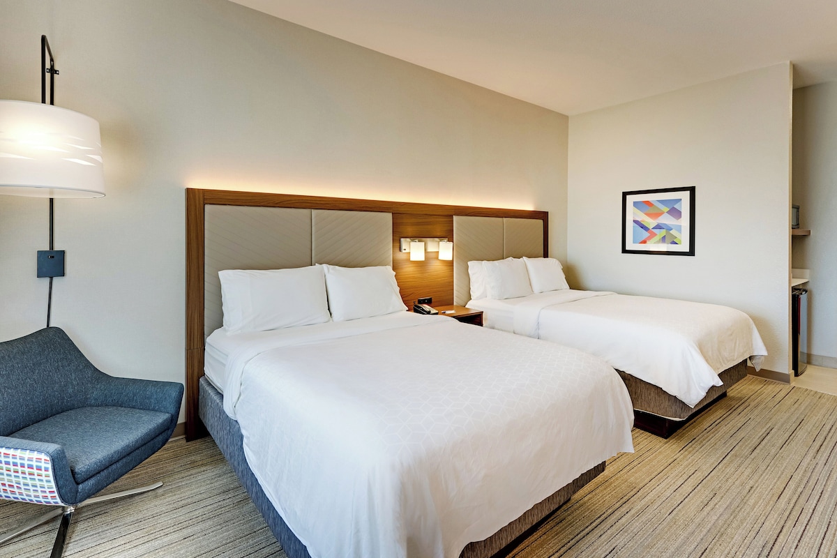 A hotel room with two beds, a desk, a chair, and a lamp.