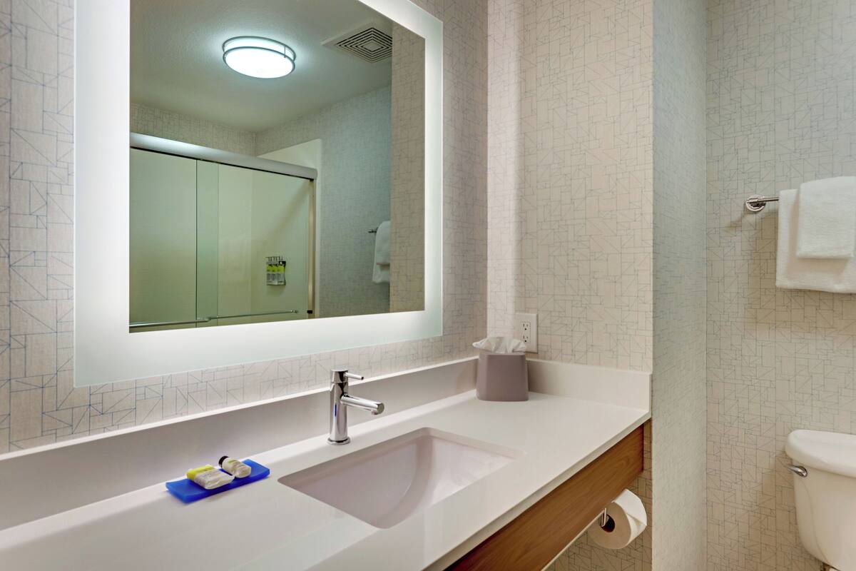 A modern bathroom with a large mirror, a white sink, a toilet, and a shower visible in the reflection.