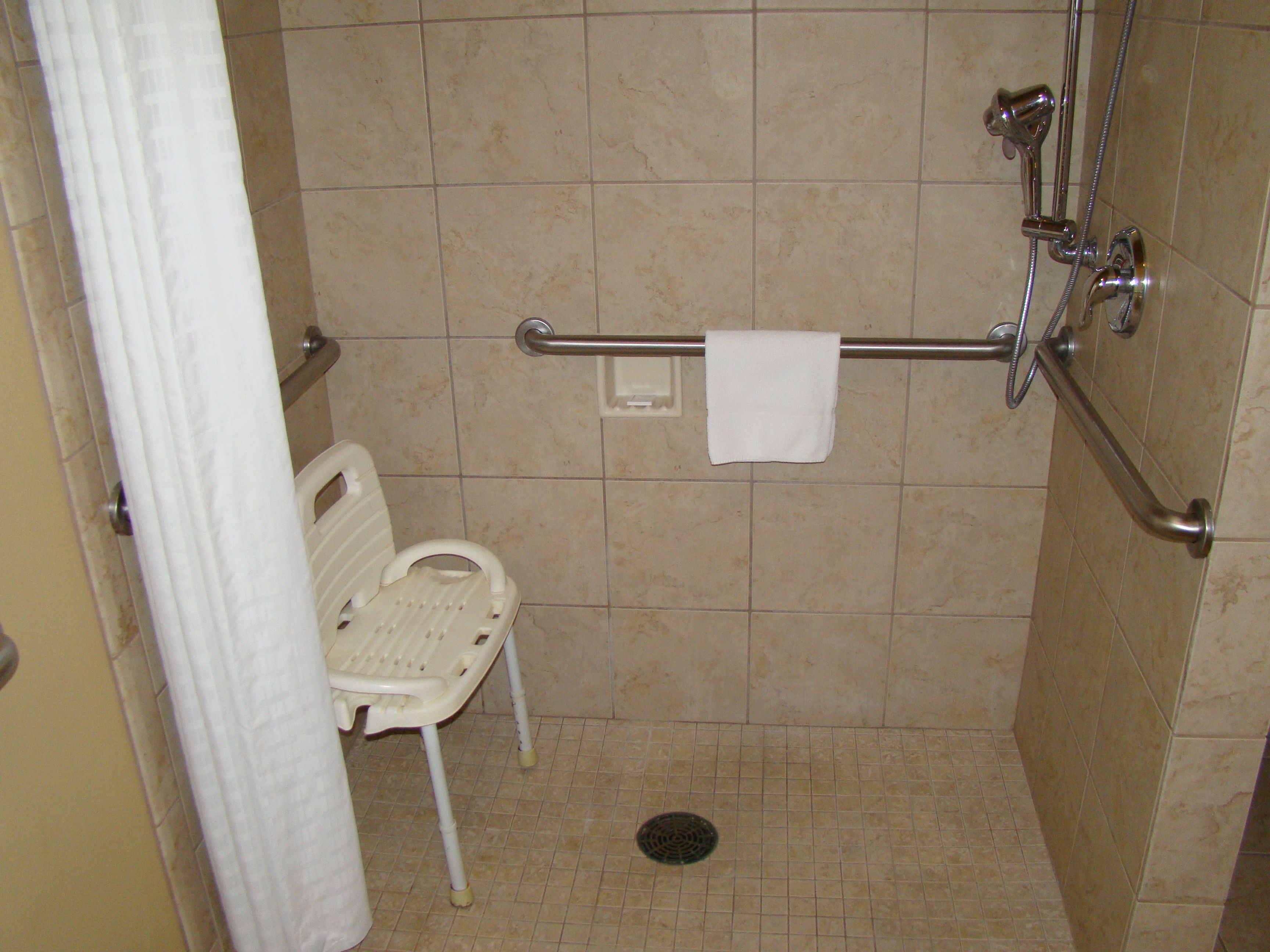 Un espacio de ducha pequeño con azulejos, una barra de apoyo, una silla de ducha blanca y una toalla colgada en la barra.