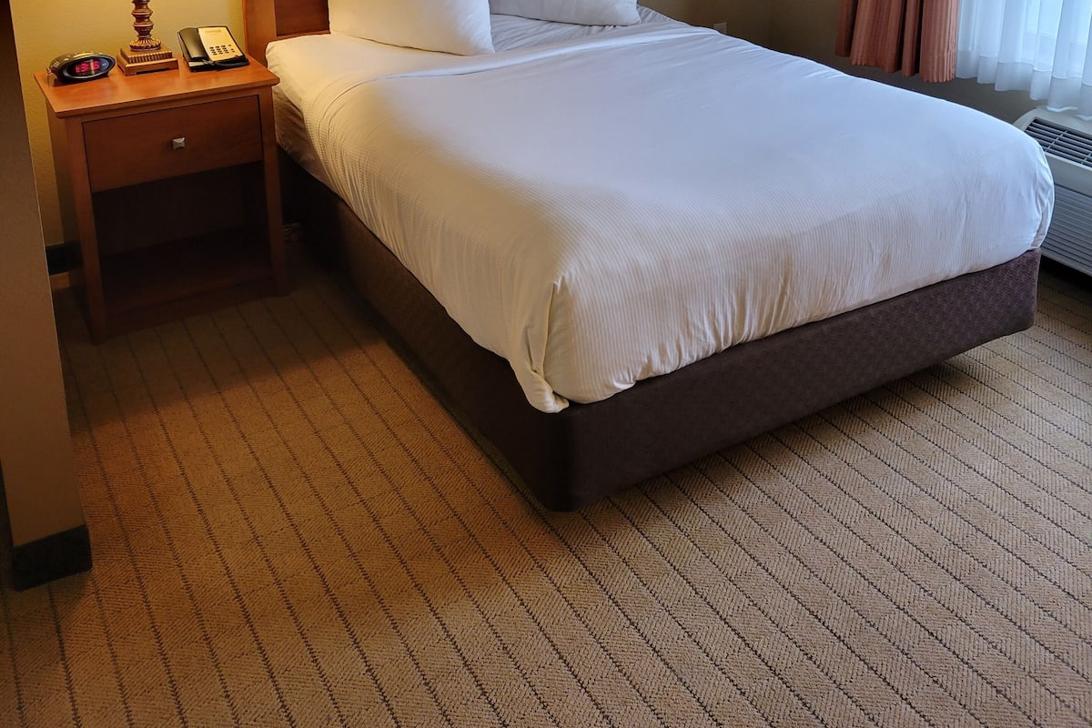 A hotel room with a large bed, a wooden headboard, a bedside lamp, a framed picture on the wall, and a window with curtains.