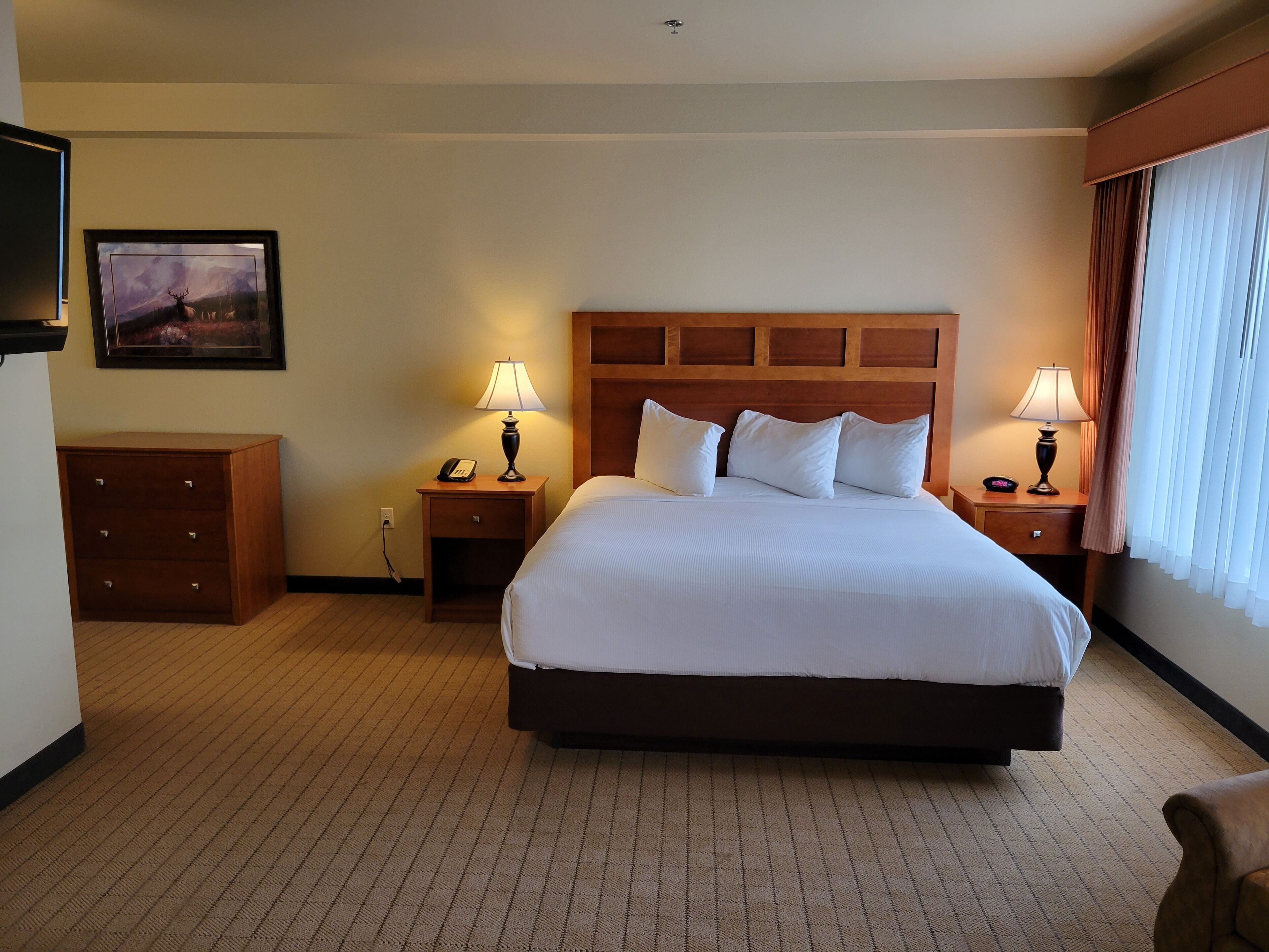 A hotel room with a large bed, two bedside tables with lamps, a wooden headboard, a small wooden dresser, and a framed picture on the wall.