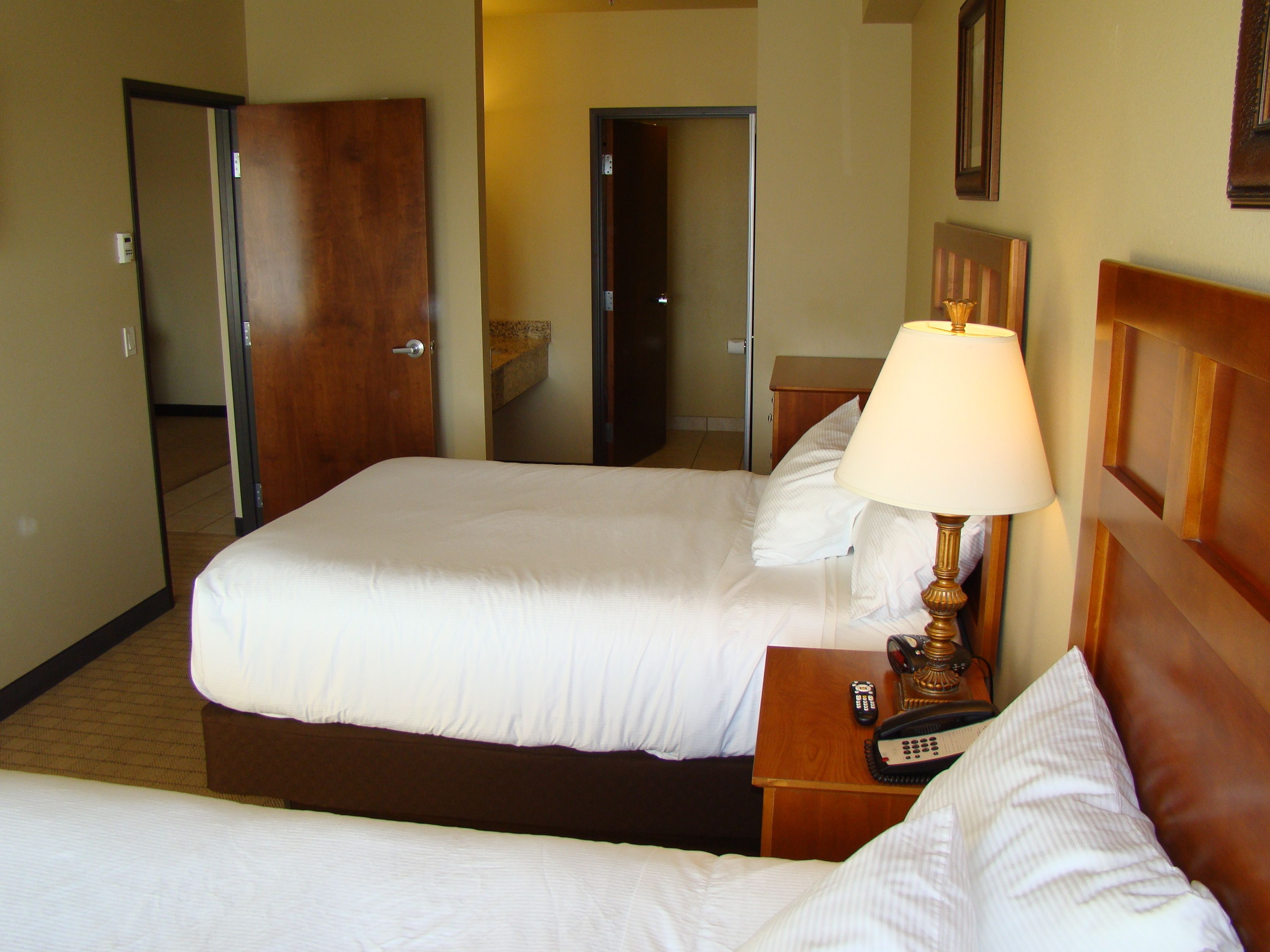 Quarto de hotel com duas camas, uma mesa de cabeceira com abajur e telefone, e uma porta que dá para outro quarto.