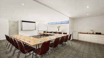 A conference room with a long table, red chairs, a projector screen, and a small kitchenette area.