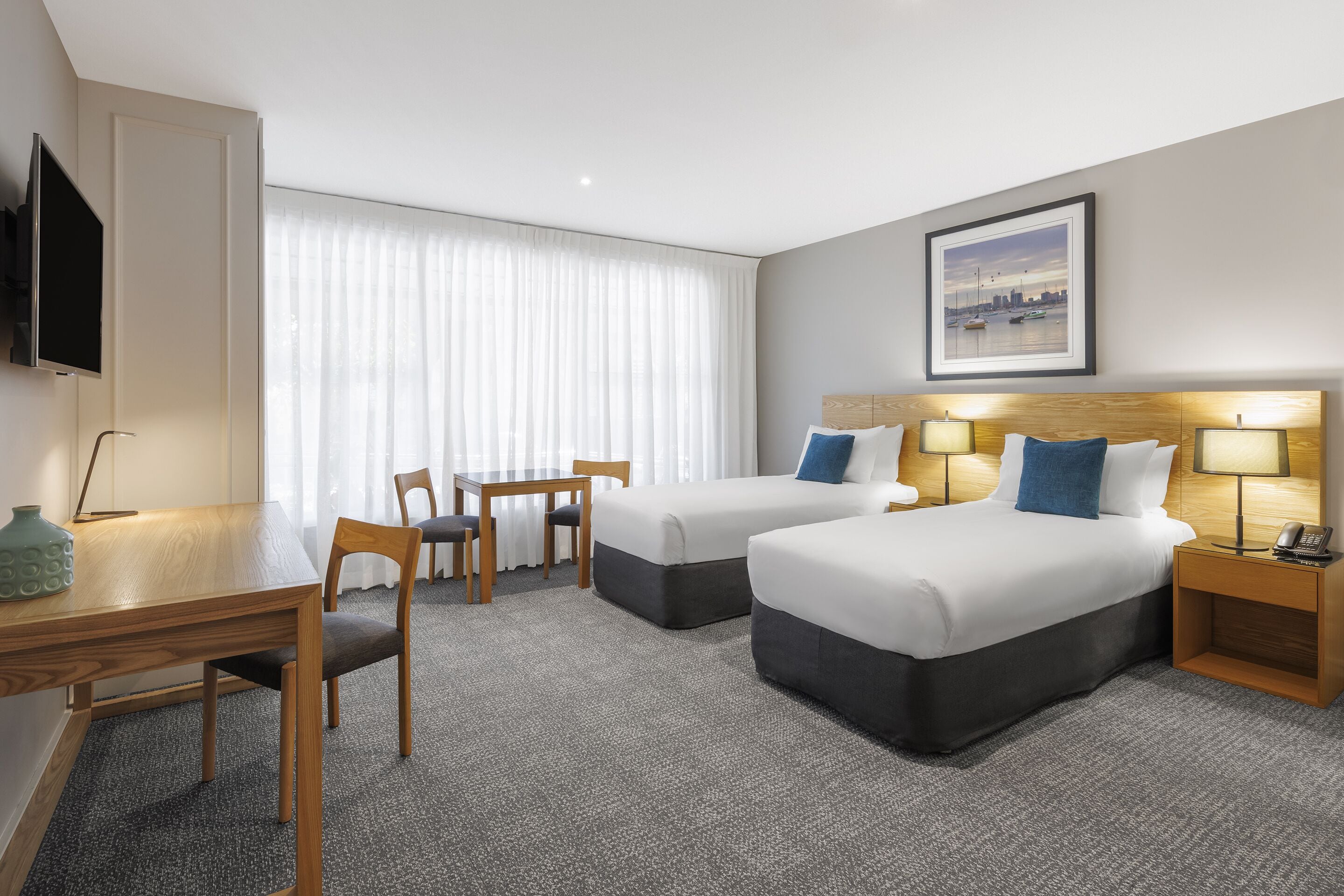 A hotel room with two beds, a desk, chairs, a TV, and a painting on the wall.
