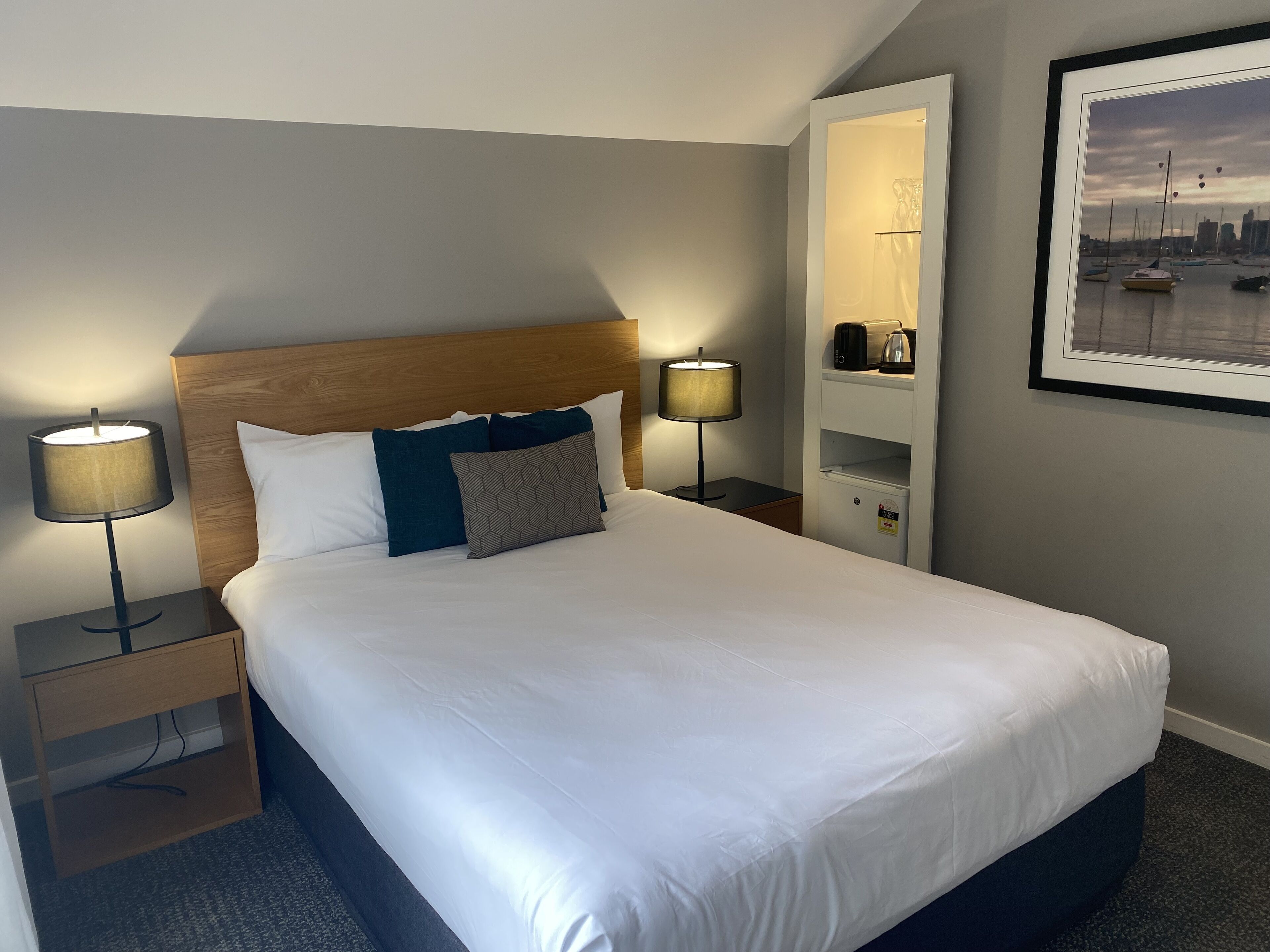 A neatly made bed with white linens and two pillows, flanked by bedside tables with lamps. A framed picture hangs on the wall to the right.