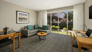 Una habitación de hotel moderna con un sofá gris, una mesa de comedor de madera, una computadora portátil y un ventanal con vistas a palmeras y un edificio.