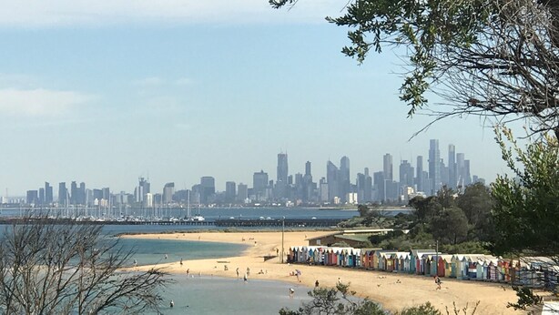 Una playa con el perfil urbano de una ciudad al fondo, un muelle y cabañas de playa.