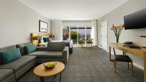 Habitación de hotel moderna con un sofá gris, una mesa de centro de madera, un escritorio con silla, un televisor de pantalla plana y un balcón con vistas a zonas verdes.