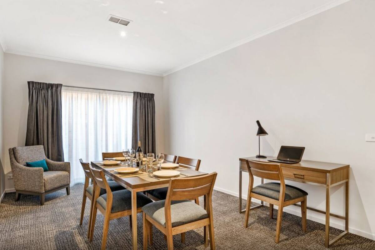 Ett hotellrum med ett matbord dukat för fyra personer, en bärbar dator på ett skrivbord, en stol med en kudde och ett fönster med gardiner.