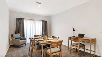 A hotel room with a dining table set for four, a laptop on a desk, a chair with a cushion, and a window with curtains.