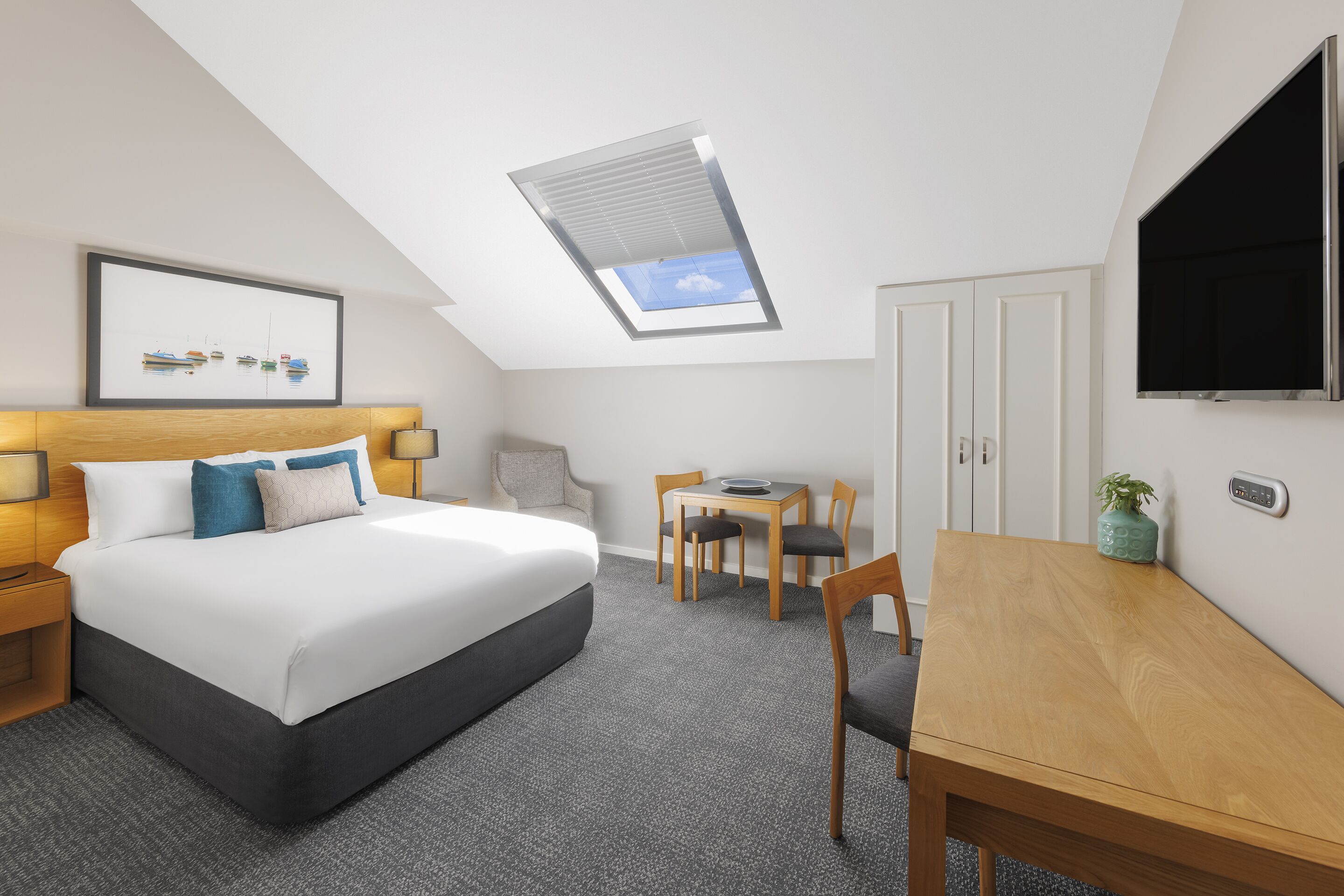 A hotel room with a bed, a desk, a chair, a TV, a framed picture, and a skylight.