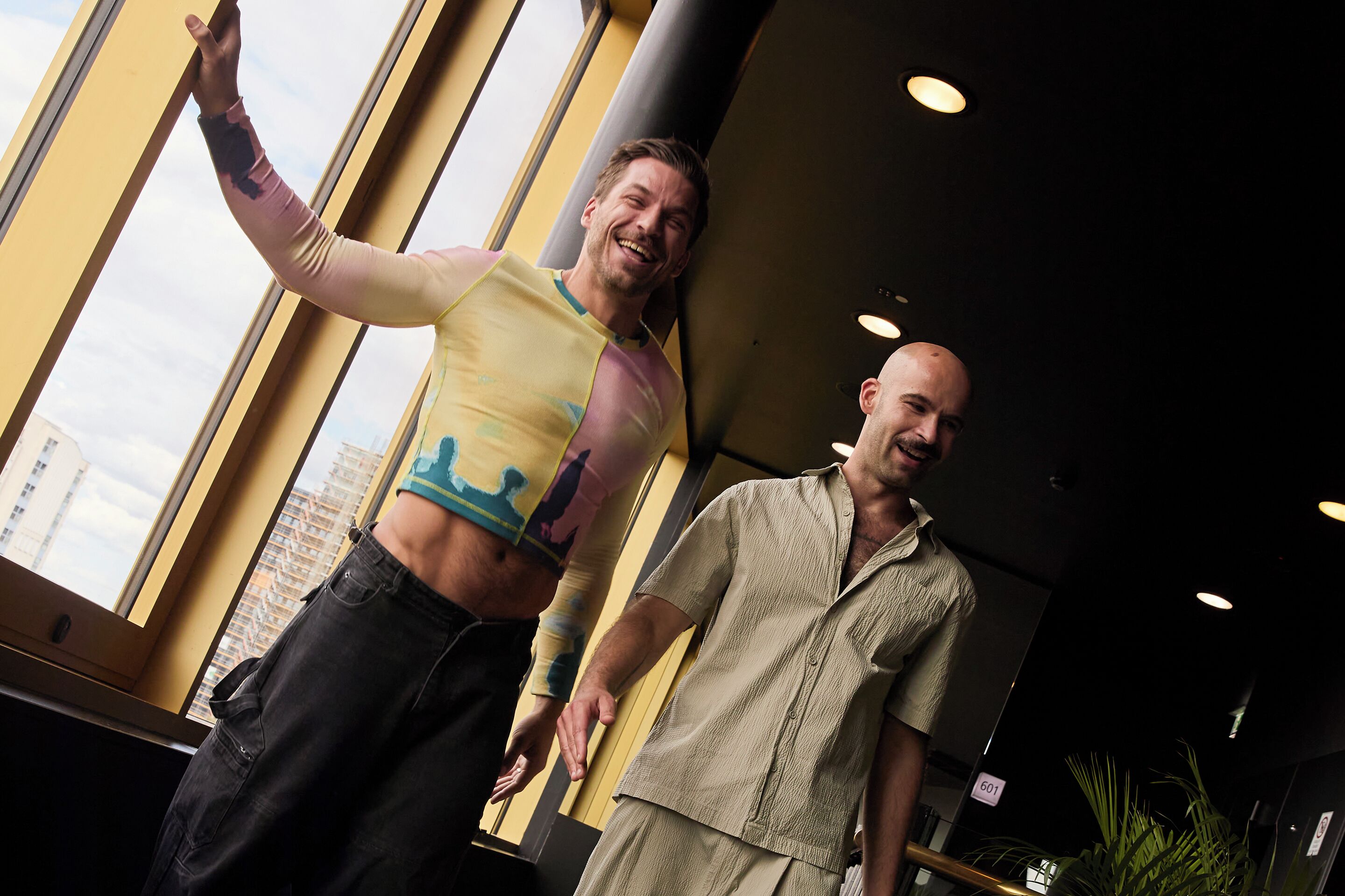 Two men standing in a room with large windows and a plant.