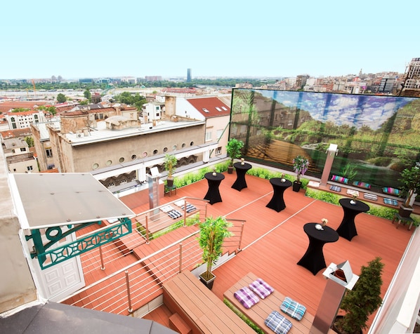 Una terraza en la azotea con asientos al aire libre, vista del paisaje urbano y una pantalla grande mostrando un paisaje.