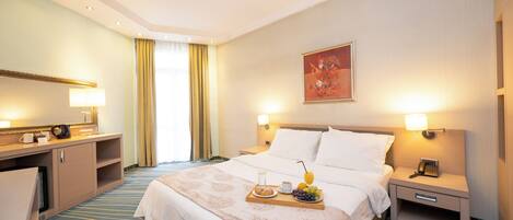 A hotel room with a large bed, bedside tables with lamps, a desk with a lamp, a window with curtains, and a painting on the wall.