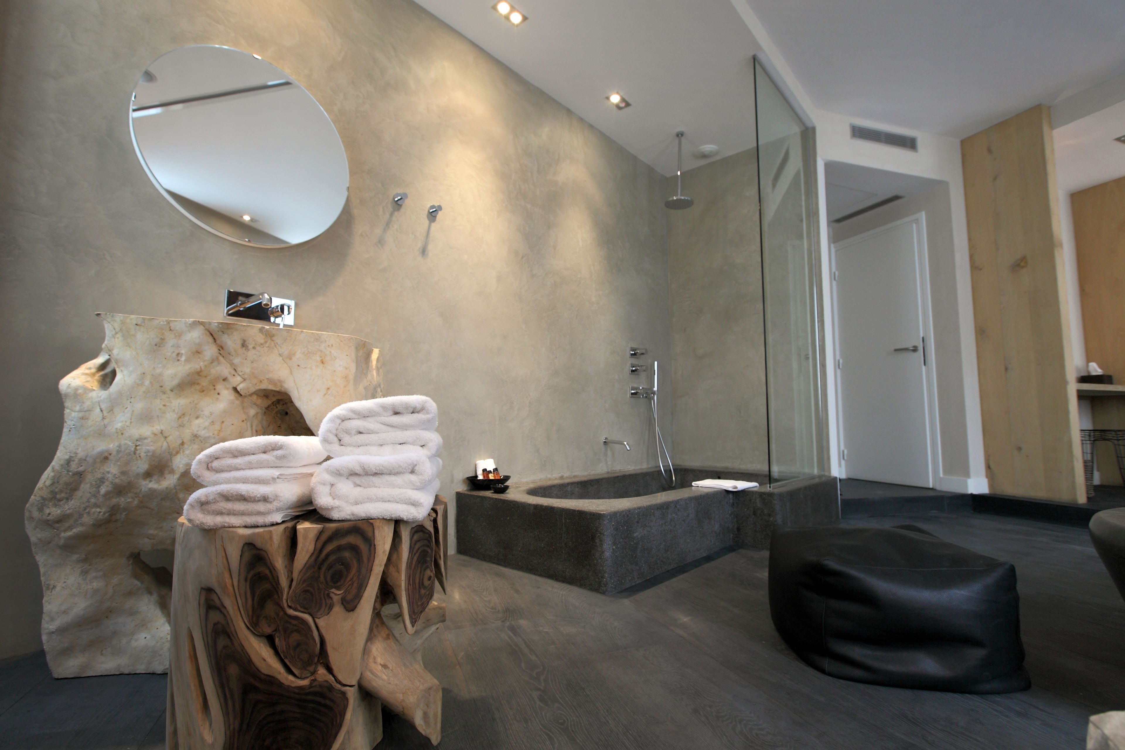 A modern bathroom with a stone sink, a large mirror, a bathtub, and a wooden stool with towels.
