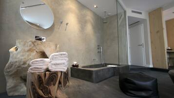 A modern bathroom with a stone sink, a large mirror, a bathtub, and a wooden stool with towels.