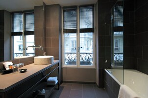 A modern bathroom with a large bathtub, a vanity with a round mirror, and a window with a view of a building.