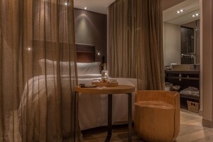 Una moderna camera d'albergo con un letto, un tavolino rotondo in legno con un vassoio di oggetti, una sedia in legno e un grande specchio che riflette la stanza.