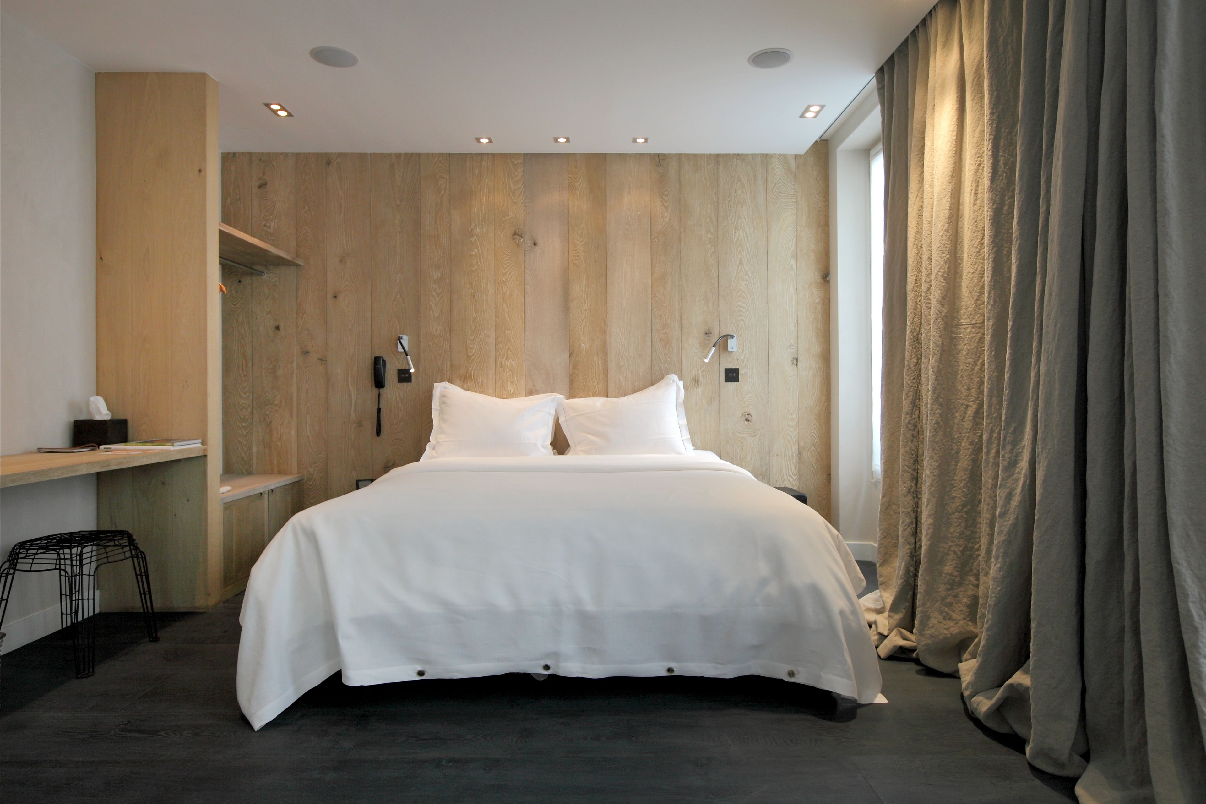 Un dormitorio moderno con una pared de madera decorativa, una cama con ropa de cama blanca, un escritorio con silla y un ventanal con cortinas.
