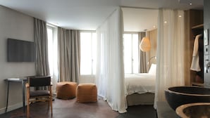 A modern hotel room with a bed, a desk, a chair, a small ottoman, a large window with curtains, and a wooden cabinet. - Hidden Hotel (Paris)
