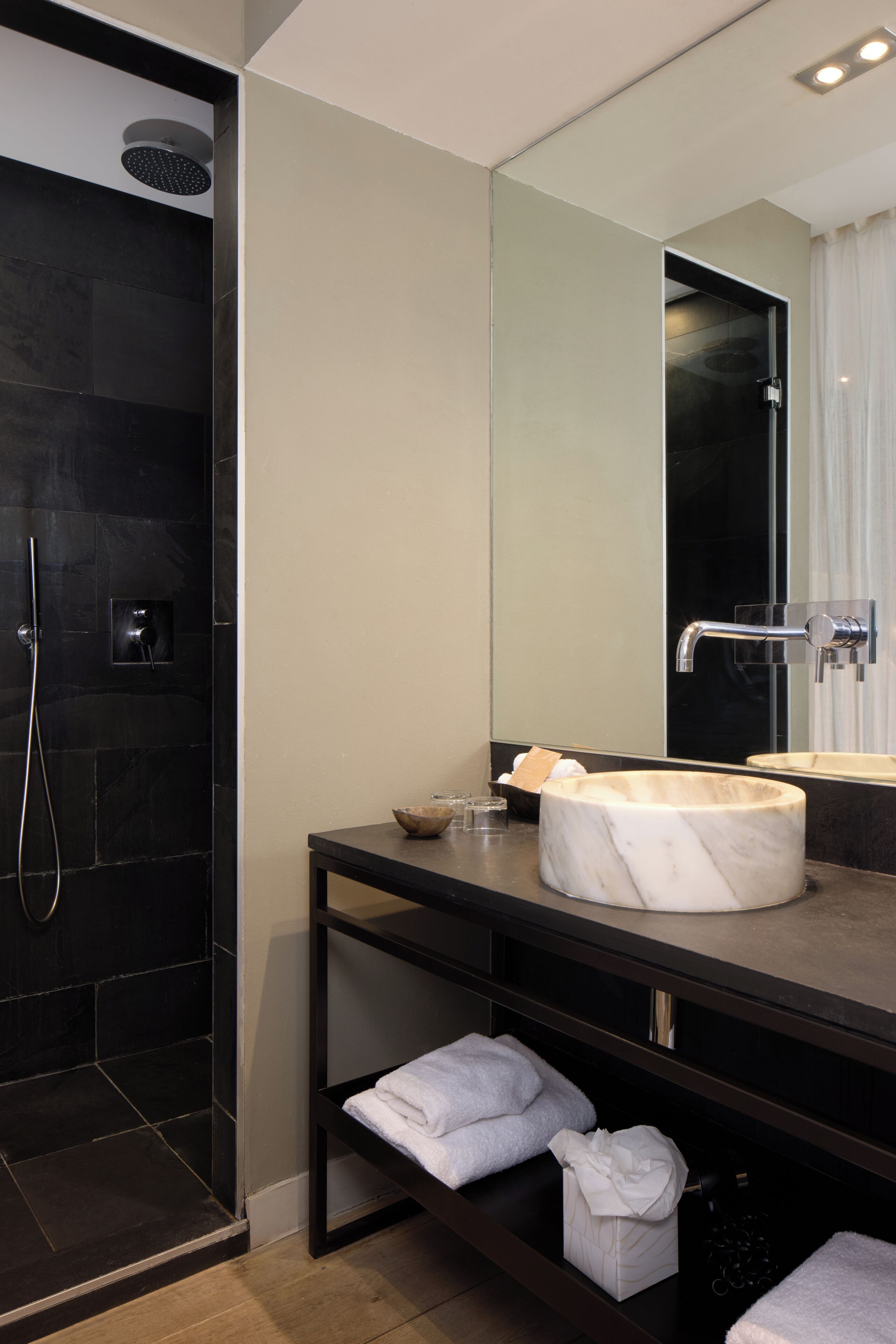 A modern bathroom with a marble sink, a large mirror, a dark shower area, and neatly folded towels on a shelf.